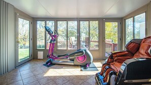 A sunlit room with a stationary bike, a massage chair, and large windows offering a view of a deck and trees. - Country  Clean  Cozy (Elizabethtown)