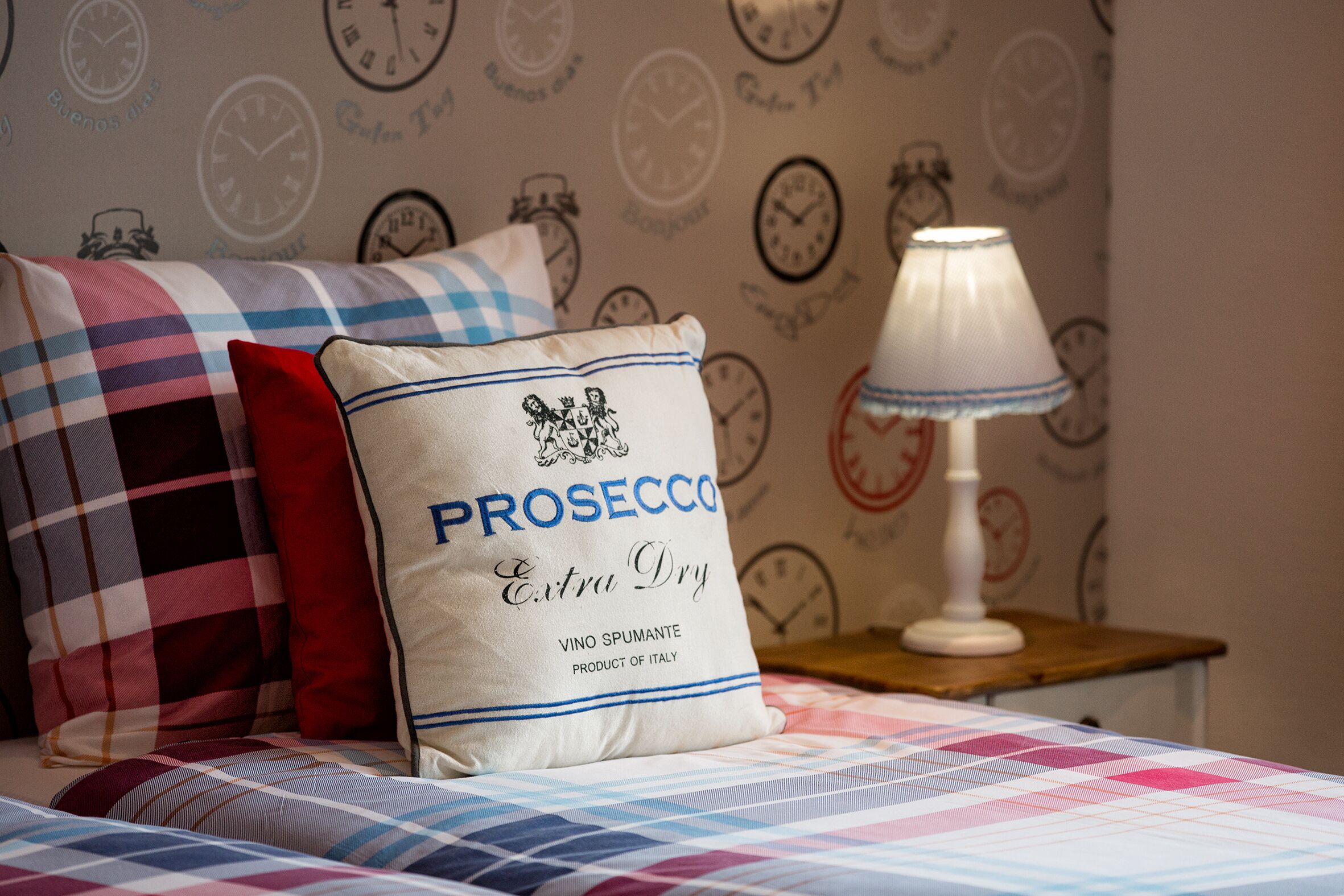 A bedroom with a bed covered in a checkered blanket, a pillow with 'PROSECCO Extra Dry' text, a bedside lamp, and a wall with clock patterns.