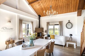 Een ruime woonkamer met een witte bank, een eettafel met stoelen, een kroonluchter en een grote klok aan de muur.