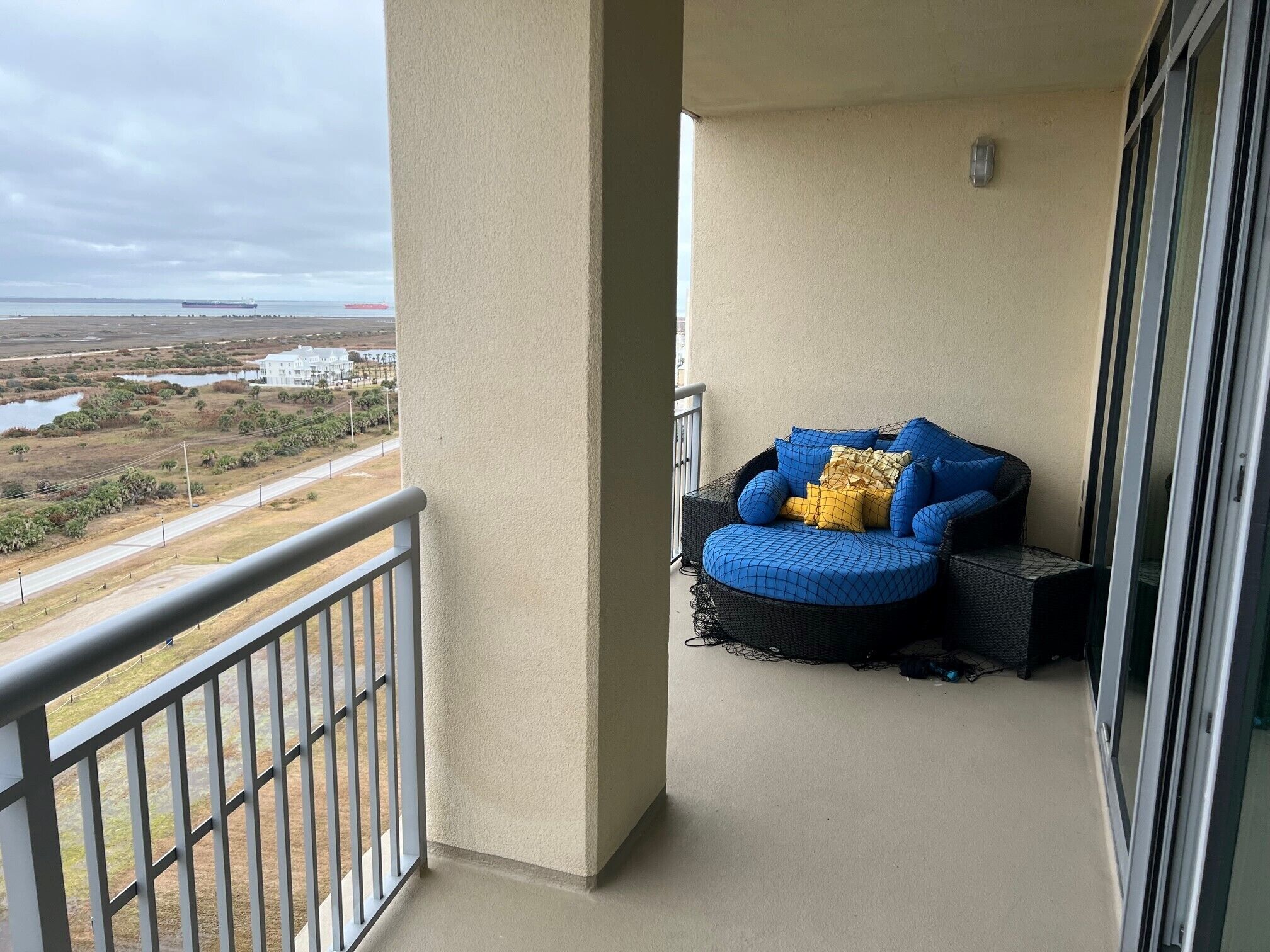 Een balkon met een zachte zithoek, uitzicht op een kustlandschap, en een gebouw in de verte.