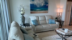 A living room with a beige sofa, a glass coffee table, and a painting of a beach scene.