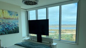 A bedroom with a large window, a flat-screen TV, a bed with a white and blue bedspread, and a painting on the wall.