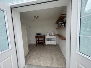 A narrow hallway with a white door, a small table with a microwave, a coffee maker, and a shelf with boxes.