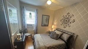 Une chambre à coucher comprenant un lit, un bureau, une chaise, un ventilateur de plafond et une fenêtre avec des rideaux.