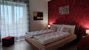 Un dormitorio con cama, mesita de noche, pared roja y ventana con cortinas traslúcidas.