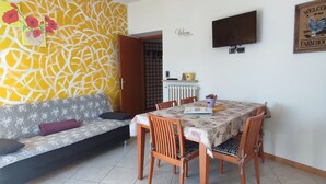 Un área de comedor con una mesa y sillas, un sofá, un televisor montado en la pared y un papel tapiz decorativo.