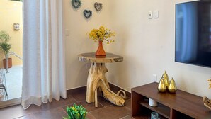 Una sala de estar con televisión, una mesa de madera con un florero, una silla de madera y un estante pequeño con adornos.