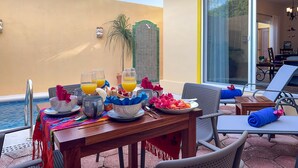 Zona de comedor junto a la piscina, con una mesa dispuesta para el desayuno, incluyendo platos, tazas y un frutero.