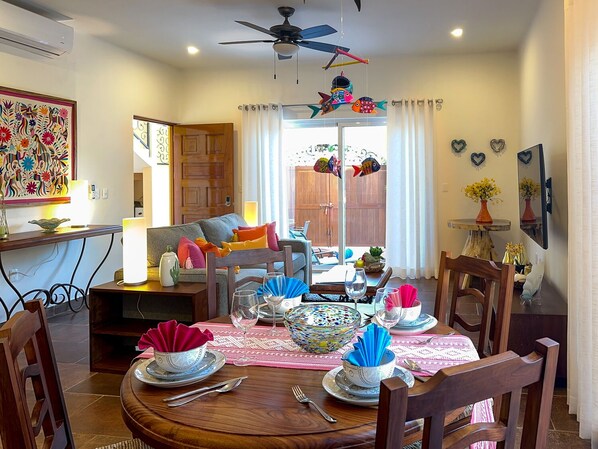 Un coin repas avec une table ronde dressée pour quatre personnes, entourée de chaises en bois. Un ventilateur de plafond se trouve au-dessus de la table, et une décoration représentant des poissons multicolores est suspendue au plafond.