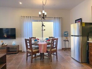 Un coin repas avec une table dressée pour quatre personnes, entourée de chaises en bois. Un lustre est suspendu au-dessus de la table, et un distributeur d’eau est visible sur la droite.