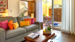 Una sala de estar con un sofá gris, cojines coloridos, una mesa de centro de madera y una puerta que da a un balcón con mobiliario exterior.