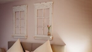 Un dormitorio con cama, almohadas, mesita de noche, lámpara y dos ventanas con contraventanas.