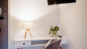Un espacio de oficina moderno con un escritorio blanco, una silla gris, un televisor montado, una lámpara y un jarrón con flores.