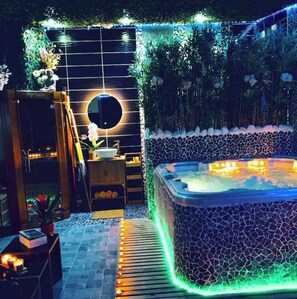 Relax in a serene oasis with a hot tub surrounded by lush greenery and decorative lighting.