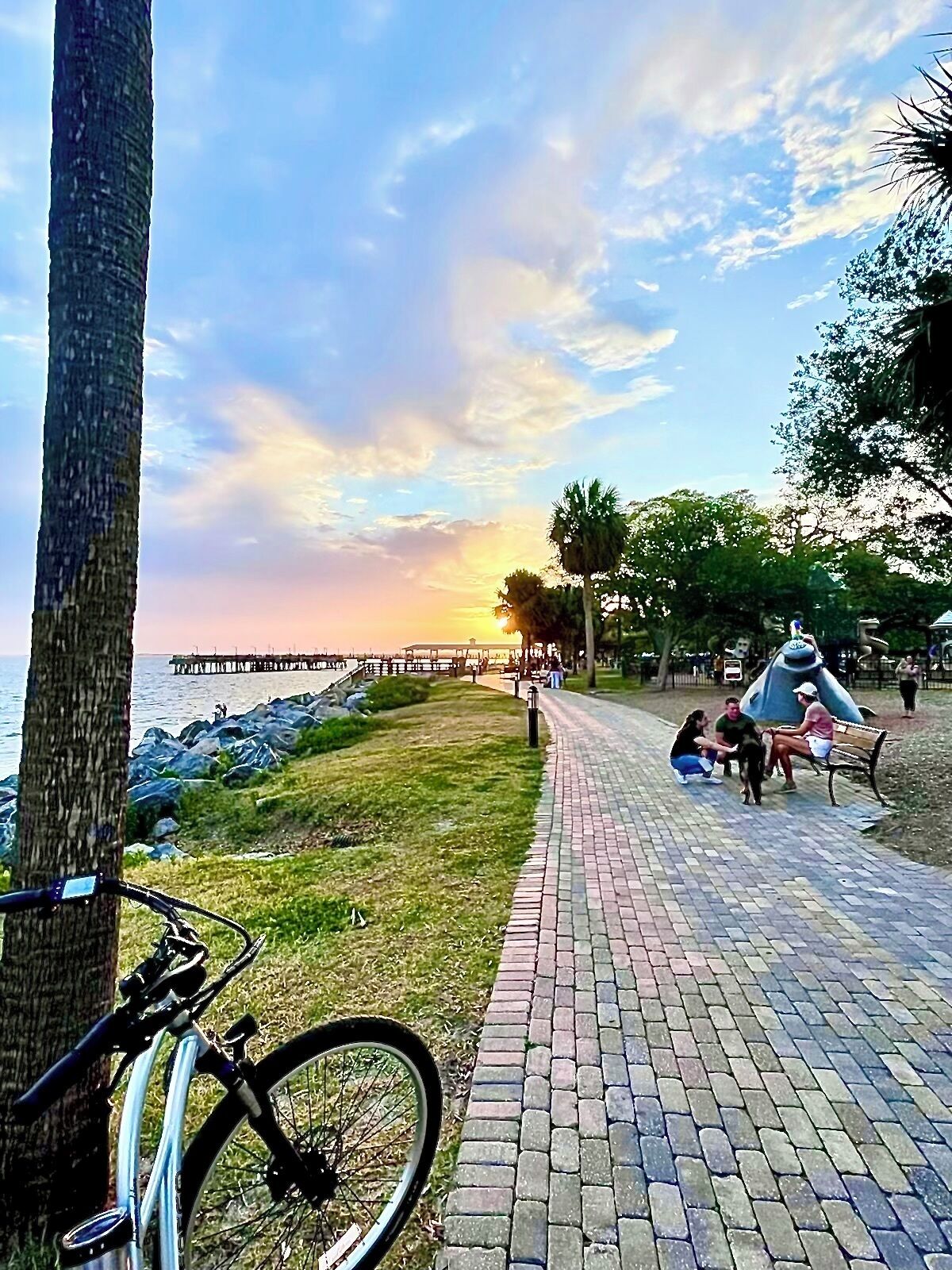 Un sendero pavimentado con una bicicleta estacionada al costado, que conduce a un parque con bancos y personas sentadas.