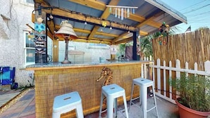Un espace bar extérieur doté d’un plafond en bois, d’une lampe de style tiki et d’une décoration représentant un hippocampe sur le mur. On y trouve deux tabourets blancs et une plante en pot.