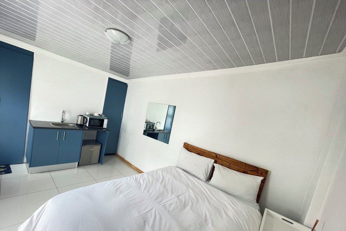 A modern bedroom with a large bed, a small kitchenette, and a bathroom visible through a mirror.