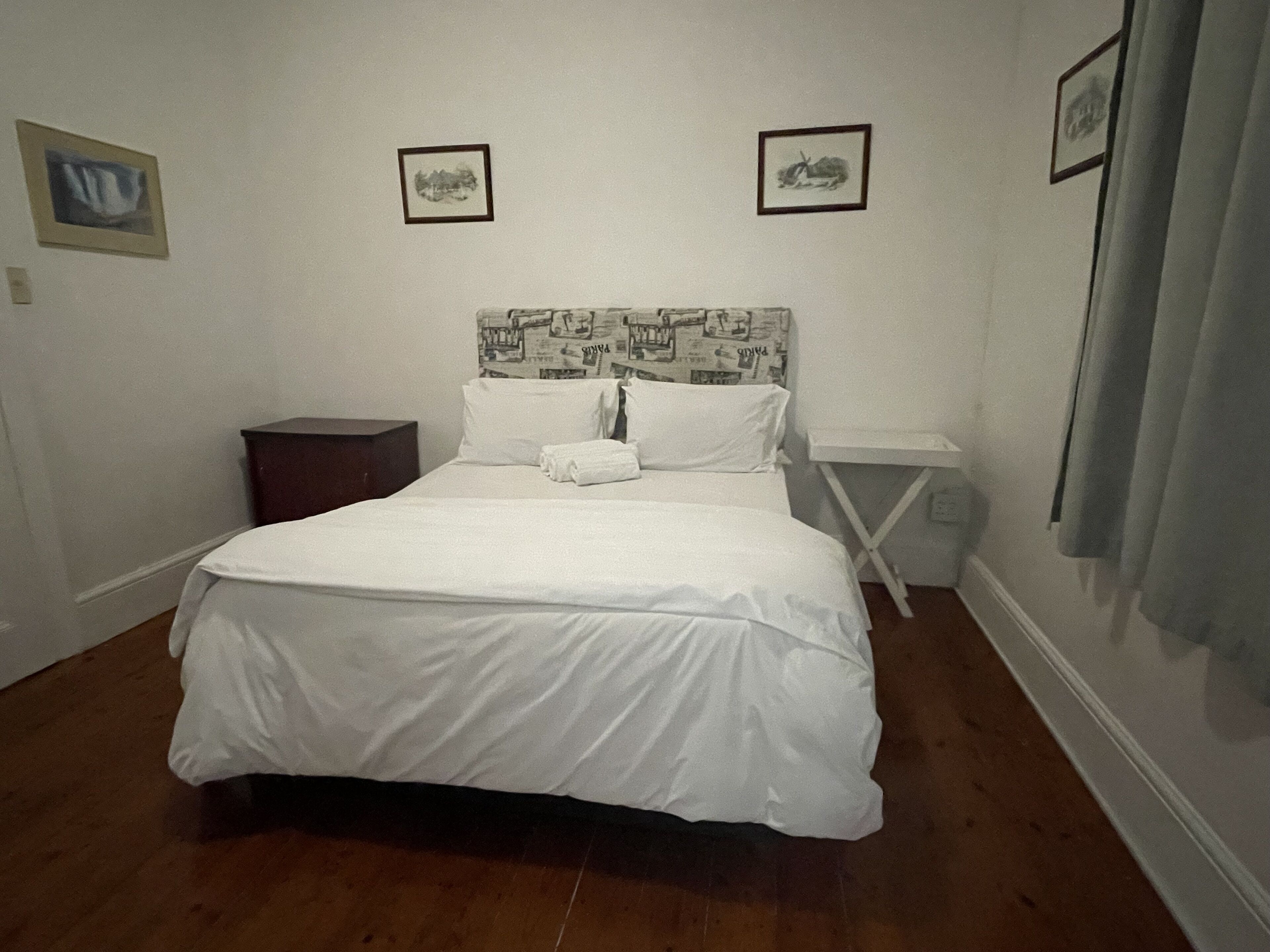Een net opgemaakt bed met witte lakens, een hoofdbord met krantenprint en twee ingelijste foto’s aan de muur.