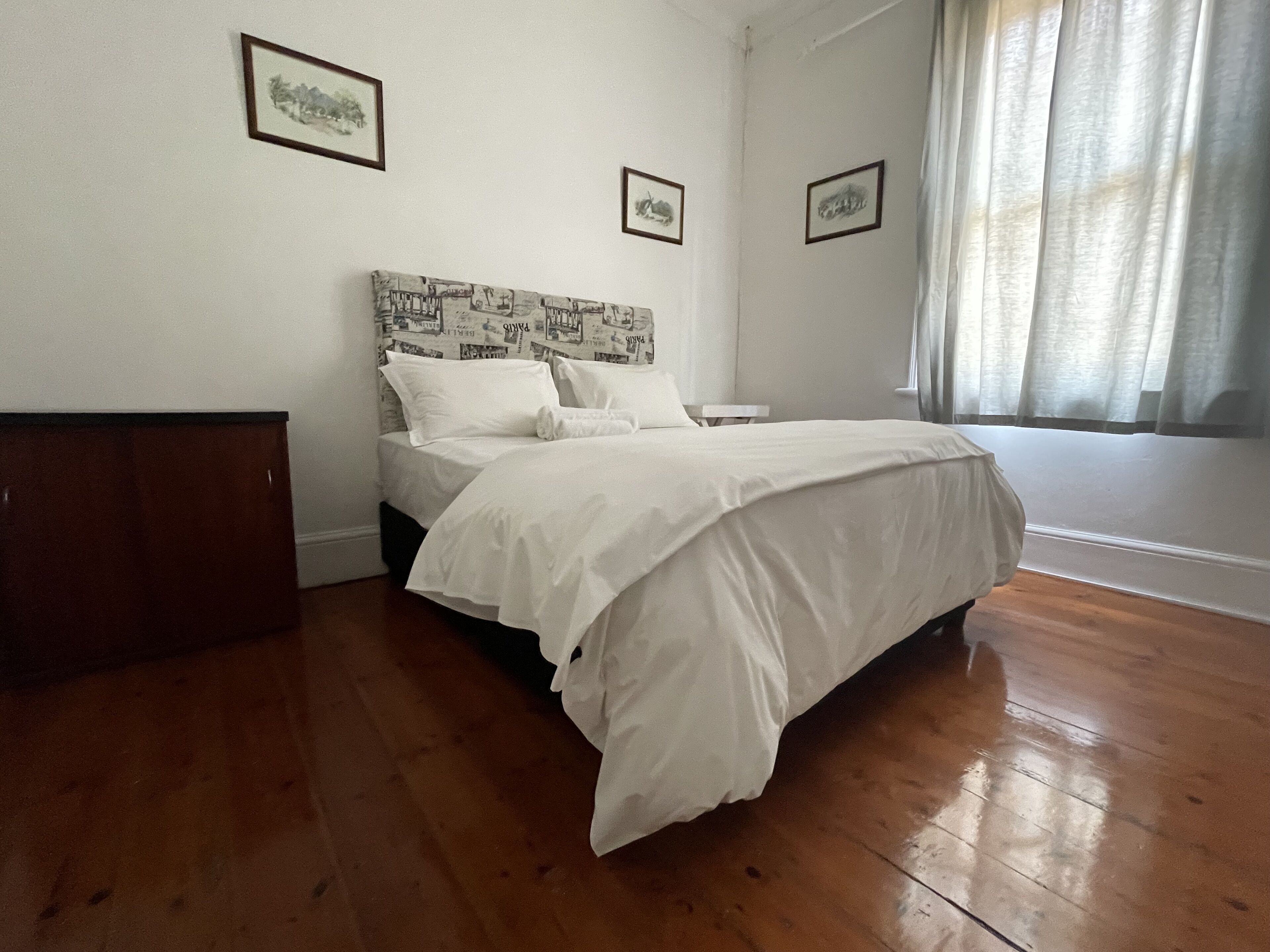 A bedroom with a wooden floor, a bed with white bedding, a nightstand, and framed pictures on the wall.