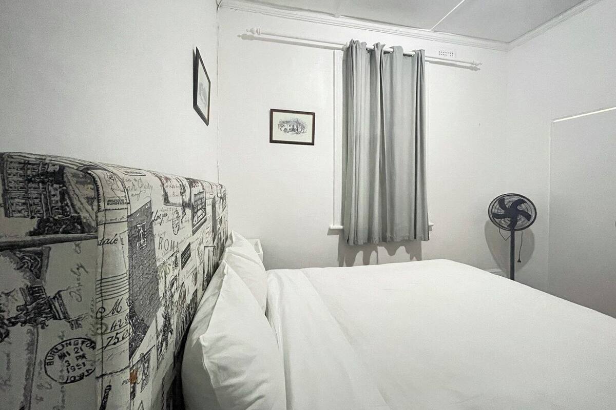 Een slaapkamer met een bed, een hoofdeinde met reis-thema's, een raam met gordijnen en een staande ventilator.