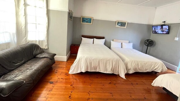 Una habitación con piso de madera, un sofá de cuero negro, dos camas individuales con ropa de cama blanca, una mesita y un televisor en la pared.