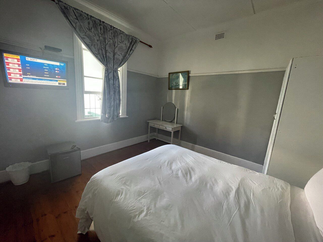 Un dormitorio con cama, una mesita de noche pequeña, un espejo, una ventana con cortinas y un televisor montado en la pared.