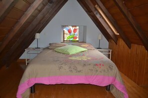 Une chambre avec un plafond en pente, un lit recouvert d’une couvre-lit à motifs floraux, une table de chevet avec une lampe, et un vitrail au-dessus du lit.