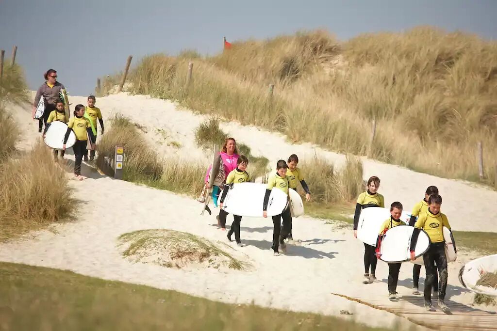 Un groupe de personnes marchant sur un sentier sablonneux, leurs planches de surf à la main, entourées de dunes et d’une végétation clairsemée.