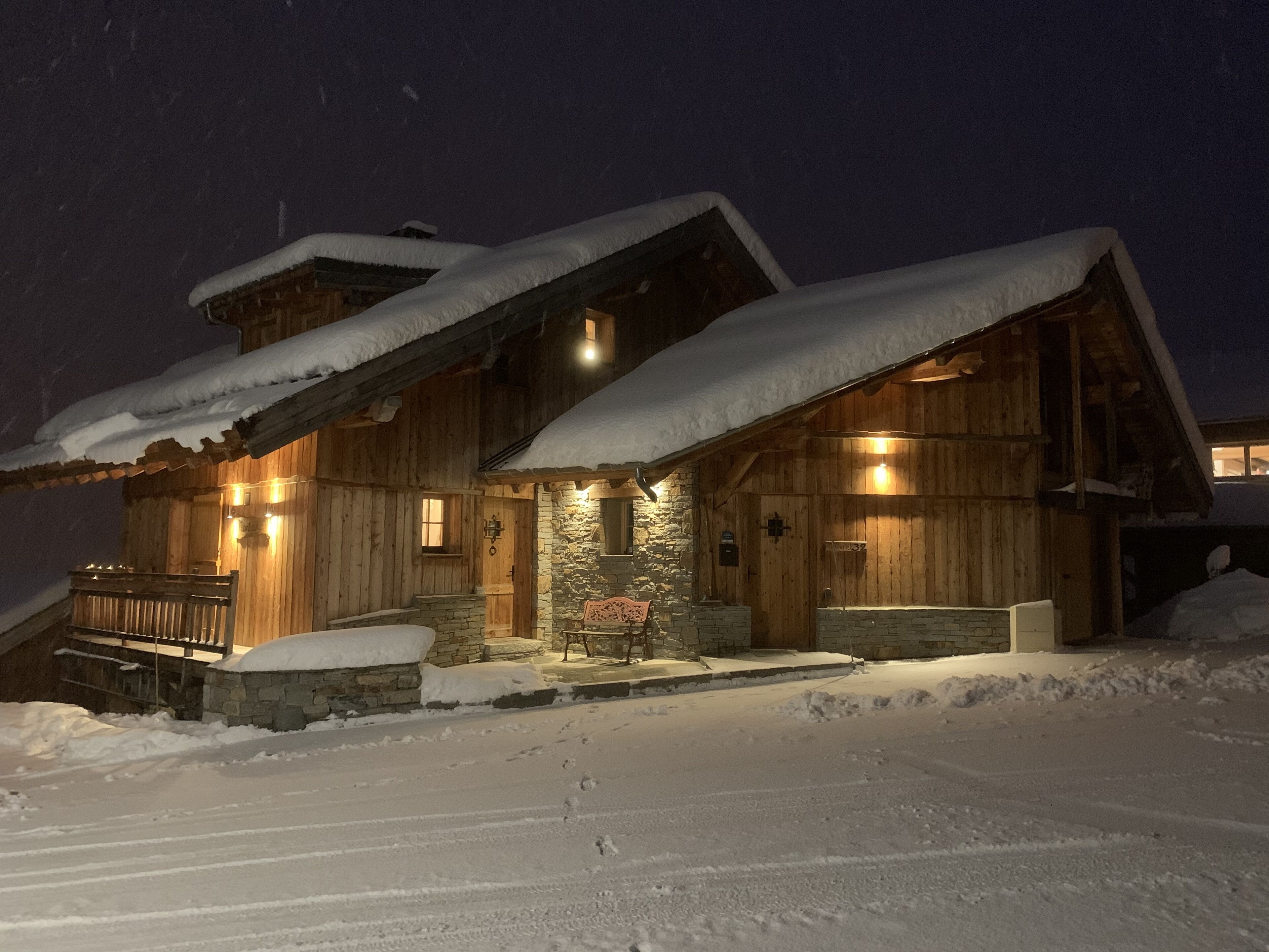 A wooden cabin with a stone foundation, snow-covered roof, and a wooden porch with a bench.