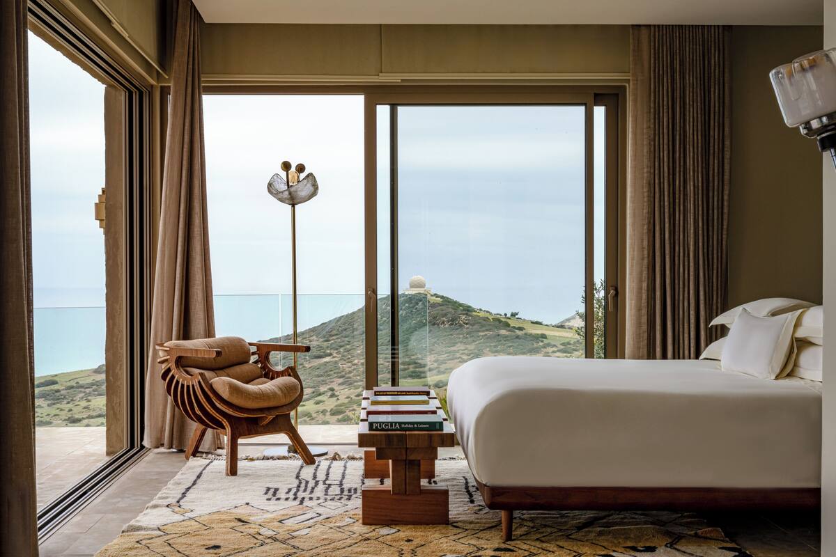 Une chambre à coucher avec un grand lit, une chaise en bois, une petite table avec des livres et une vue sur un paysage vallonné, visible à travers de grandes fenêtres.