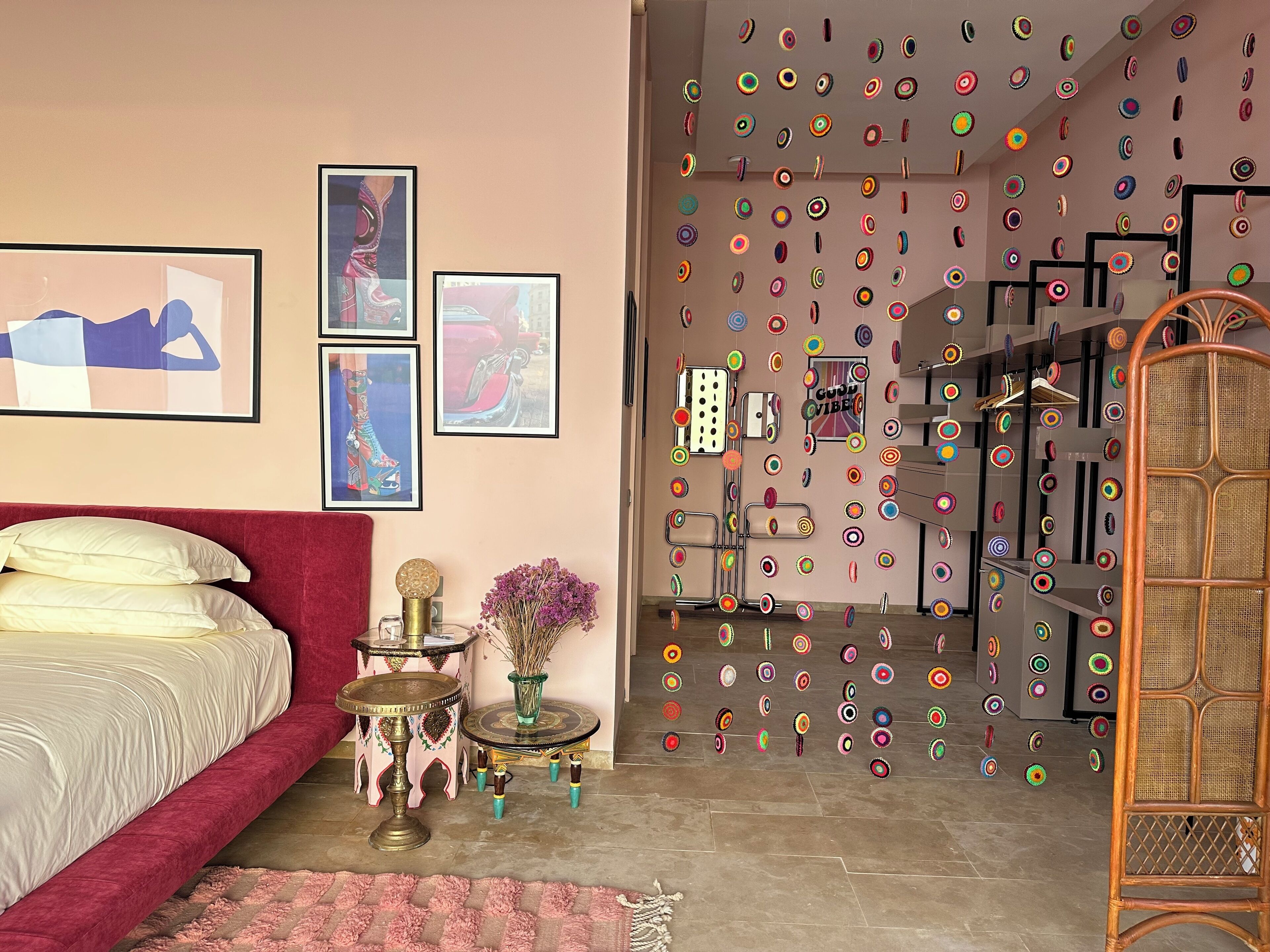 Un dormitorio con una cama roja, una mesita redonda pequeña con un objeto decorativo, un florero con flores y una pared con adornos circulares de colores.