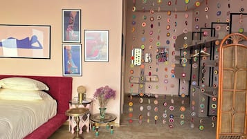 Un dormitorio con una cama roja, una mesita redonda pequeña con un objeto decorativo, un florero con flores y una pared con adornos circulares de colores.