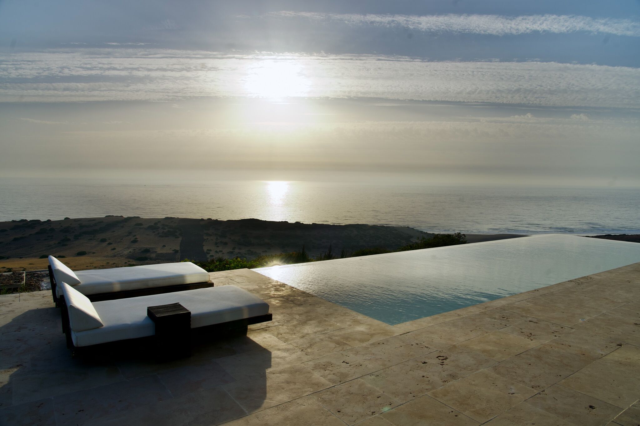A infinity pool with lounge chairs, a view of the sea, and a mountainous landscape.