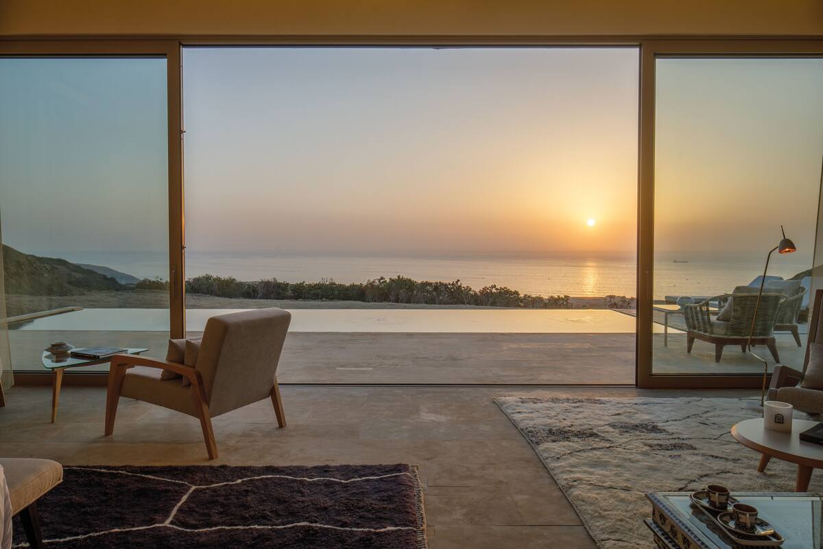 Un salon moderne doté d’une grande porte vitrée donnant sur un balcon avec vue sur une plage au coucher du soleil.