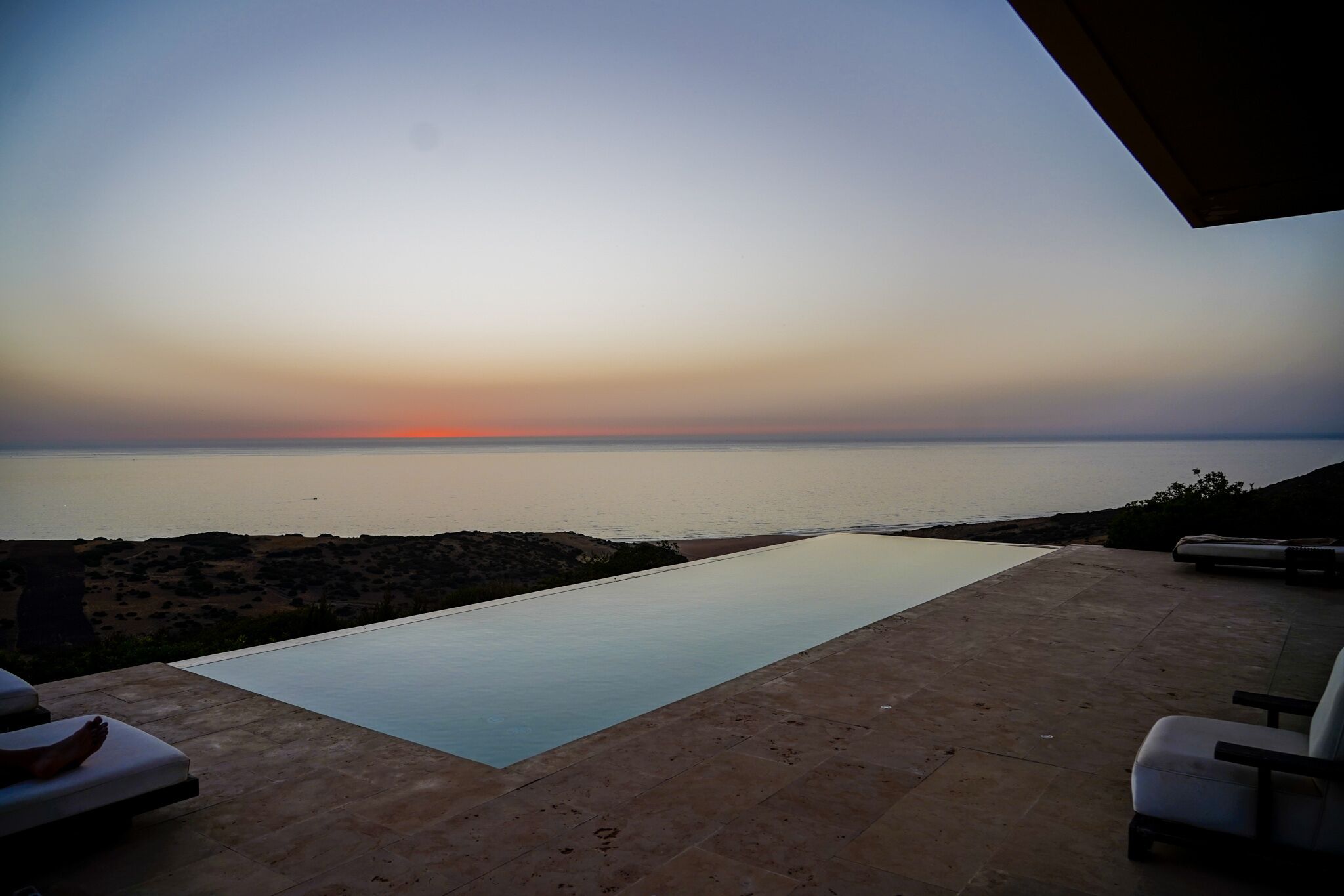 Una piscina infinita con vistas a la puesta de sol sobre el mar, mobiliario exterior y un paisaje rocoso.