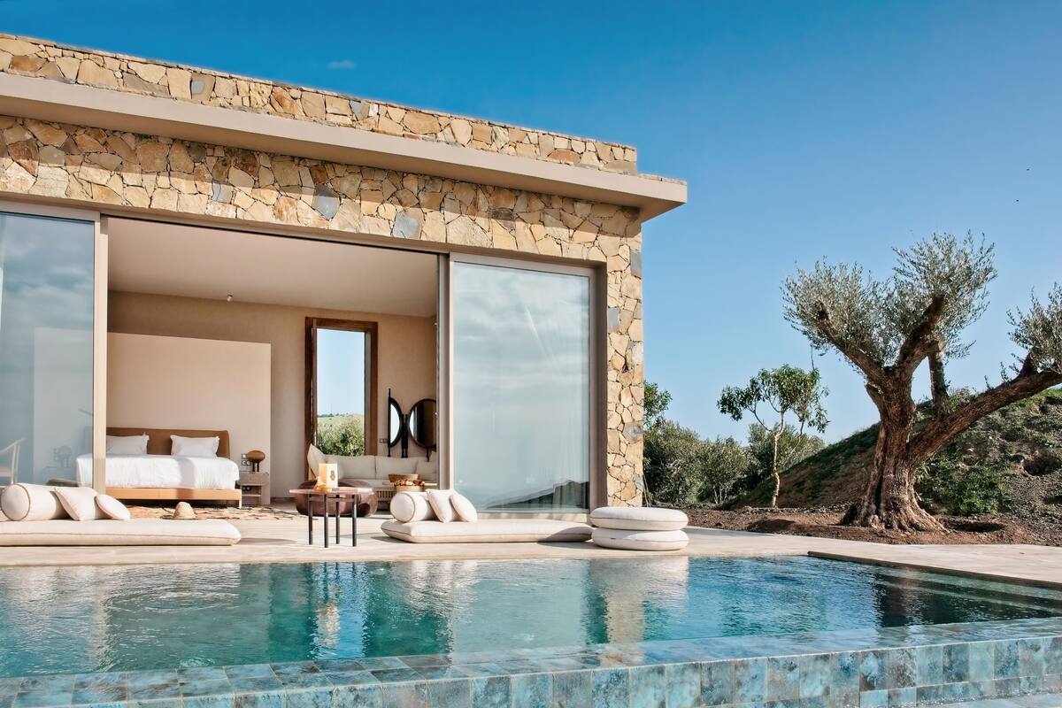 Une villa moderne avec une façade en pierre, une piscine et une chambre dotée d’un grand lit et d’un coin salon.