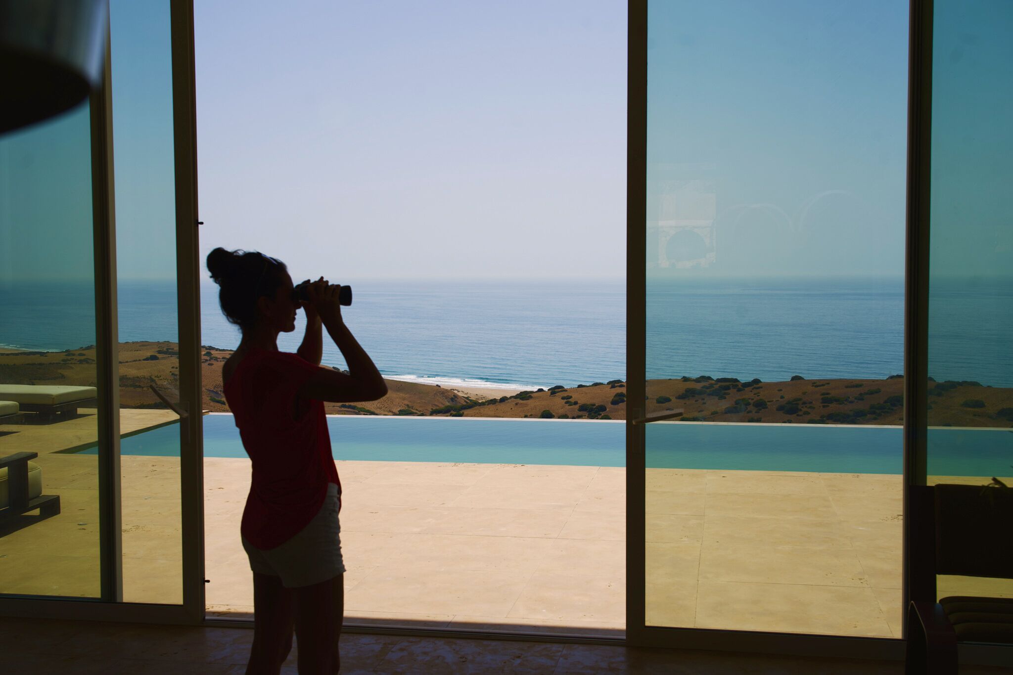 A person standing by a large window overlooking a pool and ocean view.