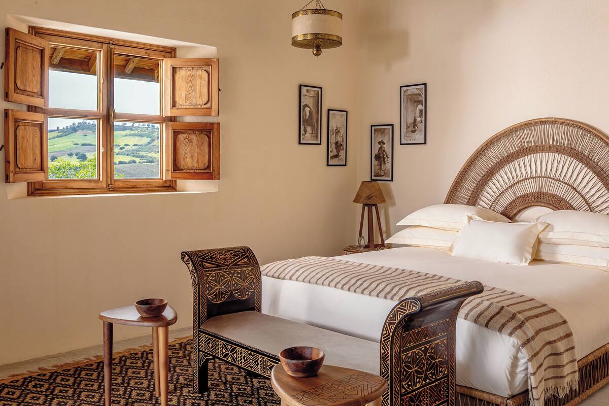Une chambre avec un grand lit, une petite table sur laquelle se trouve un bol, une chaise et une fenêtre donnant sur un paysage verdoyant.