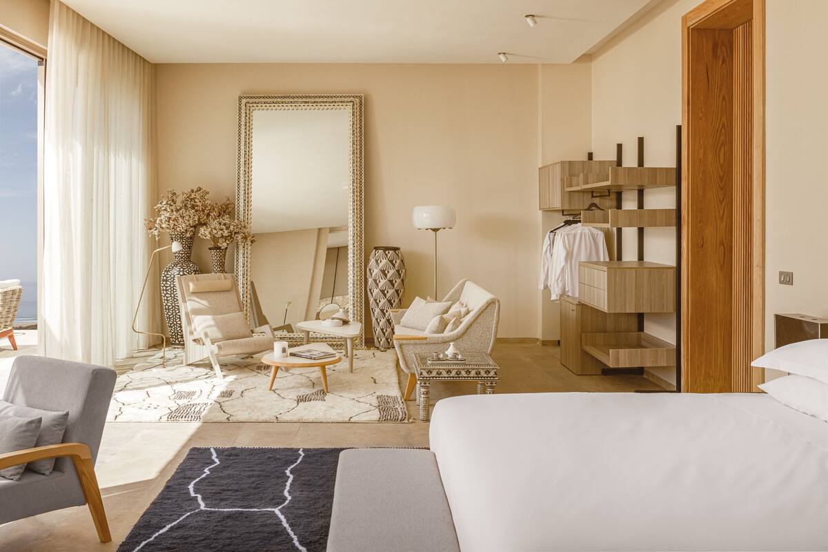 Une chambre d’hôtel avec un lit, deux fauteuils, une petite table, un miroir, une lampe, une armoire et une grande fenêtre avec des rideaux.