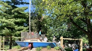 Un parque infantil con un tobogán azul y columpios, rodeado de árboles y un sendero de tierra.