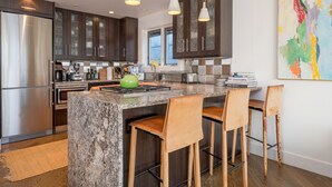 A modern kitchen with dark wood cabinets, a stainless steel refrigerator, a granite countertop, and a green kettle. There are three bar stools and a colorful abstract painting on the wall. - Sun Valley, Idaho / Downtown Ketchum Penthouse / Views, Ski, Sun, Prime Location (Ketchum)
