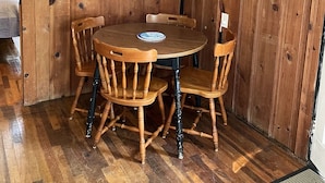 A wooden-paneled room with a round table and four chairs, a television mounted on the wall, and a framed picture of a ship. - Adorable Cape Cottage Near Kingsbury Beach (Eastham)