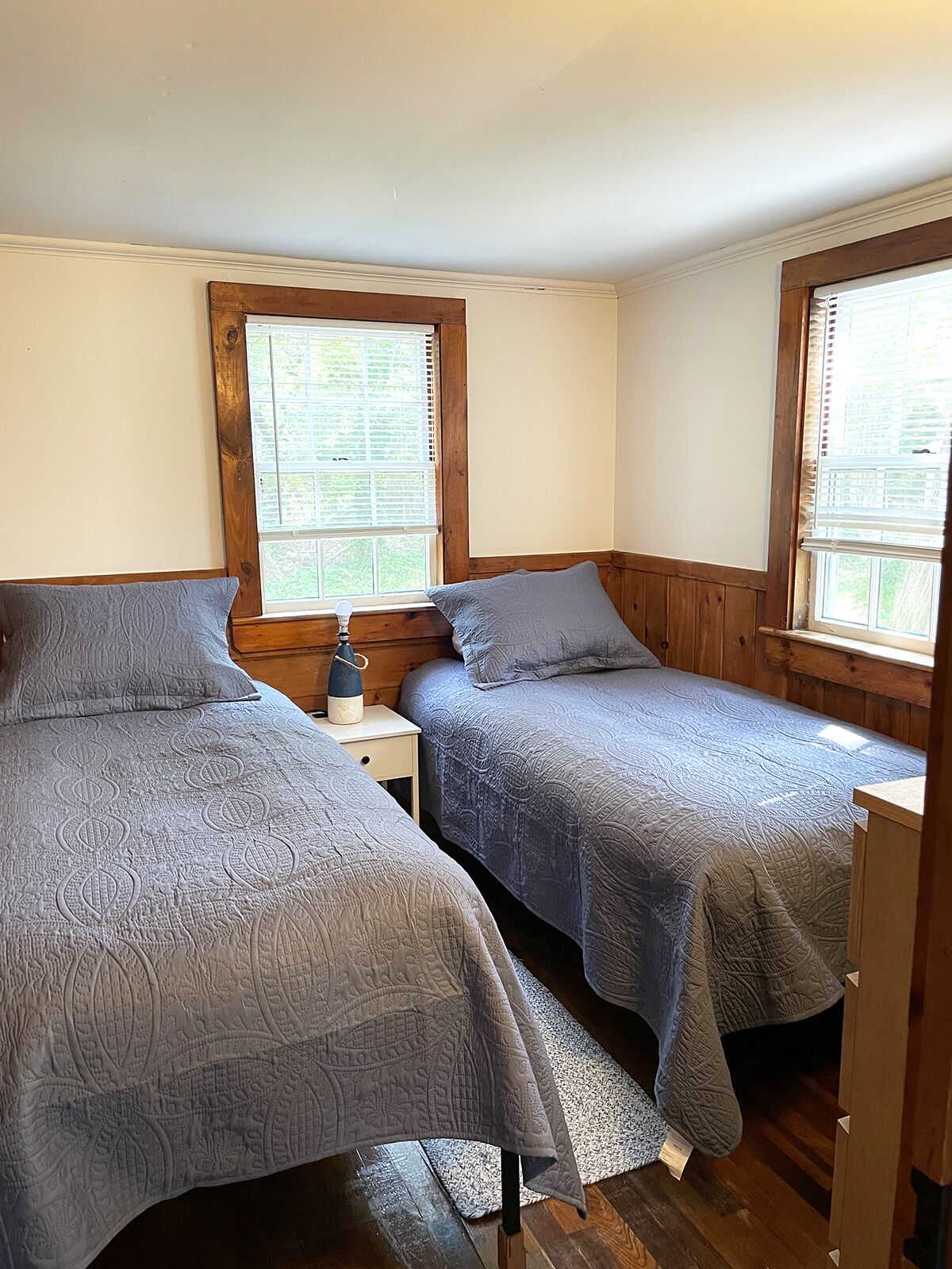 Una habitación con dos camas individuales, cabeceros de madera, una mesita de noche pequeña con lámpara y una ventana con persianas.