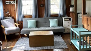 A cozy living room with a sofa, armchairs, a coffee table, and a kitchen area with cabinets and a stove. - Adorable Cape Cottage Near Kingsbury Beach (Eastham)