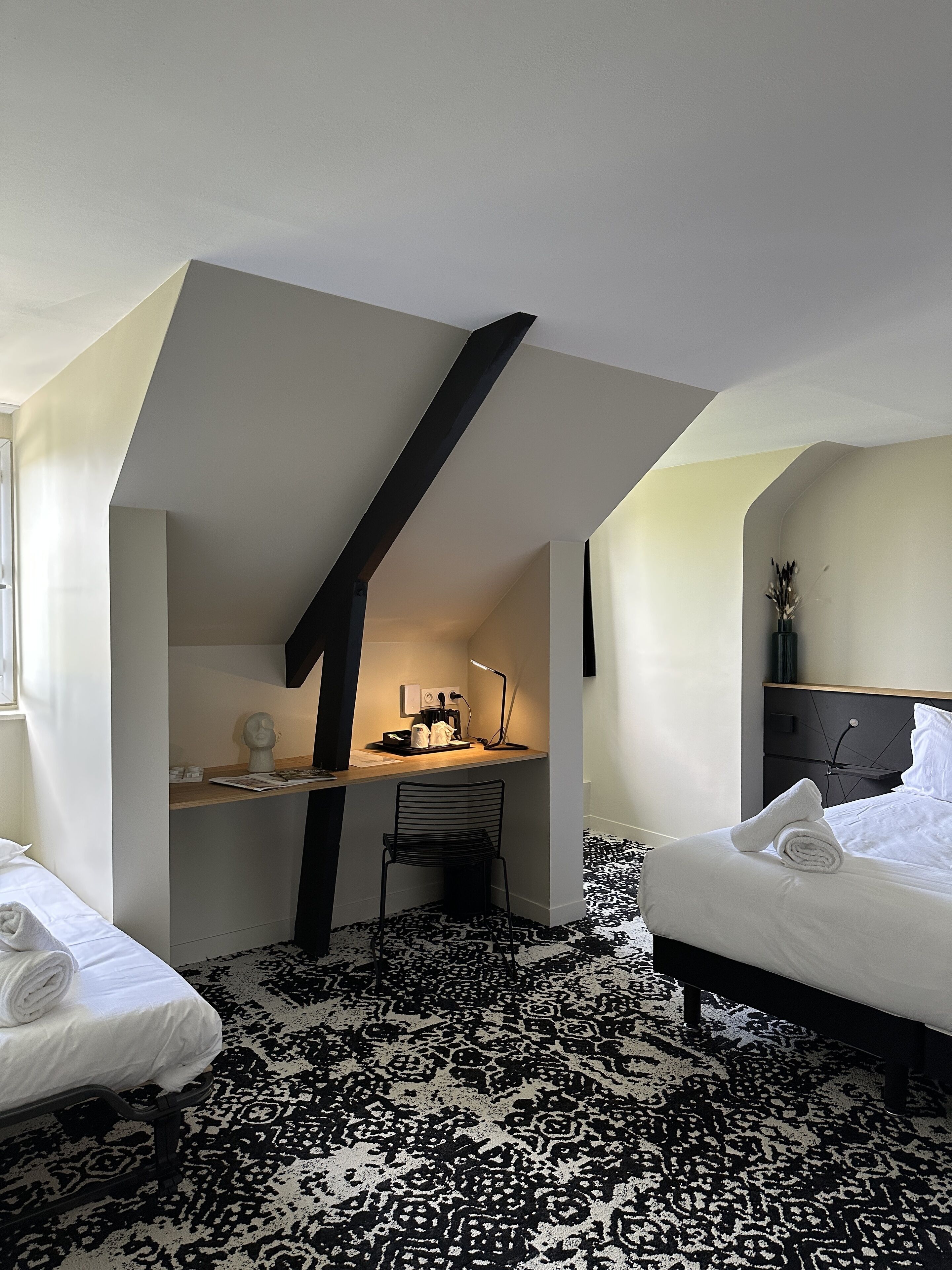 Une chambre avec un plafond en pente, un bureau avec une chaise, deux lits et une moquette à motifs.