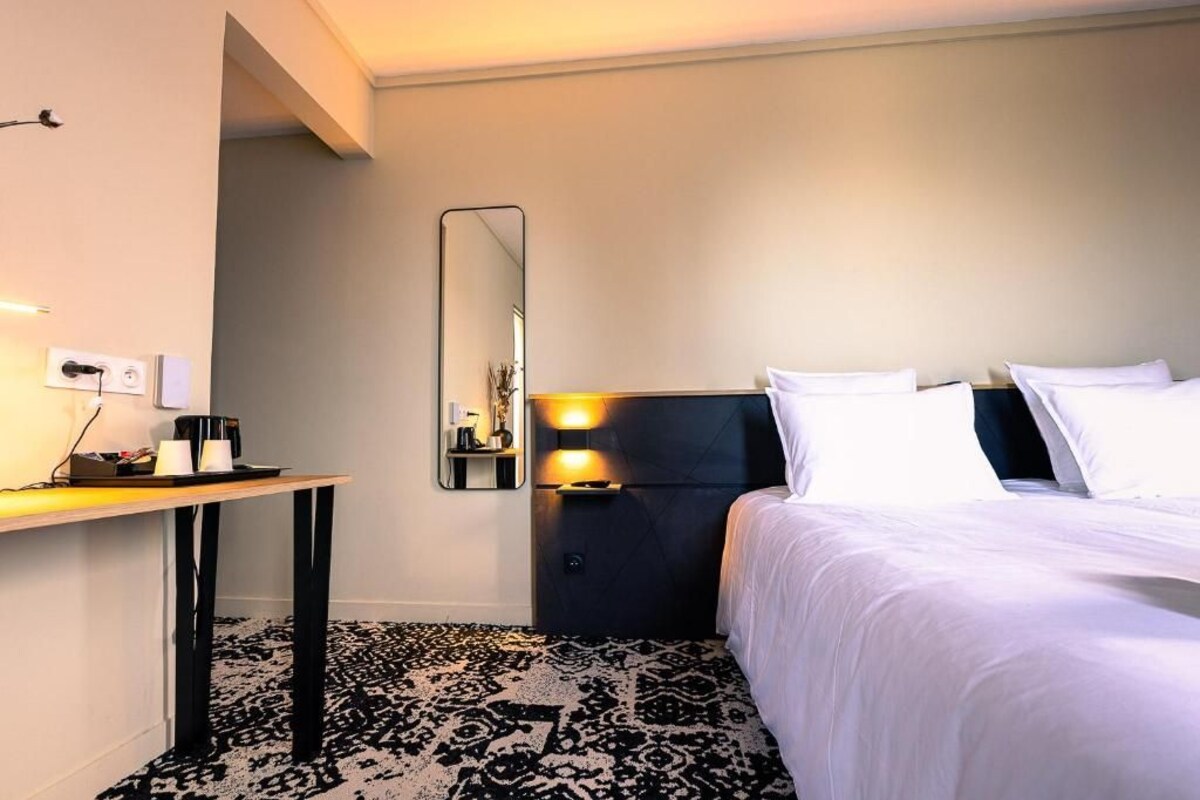 A hotel room with a bed, bedside table, mirror, and a patterned carpet.