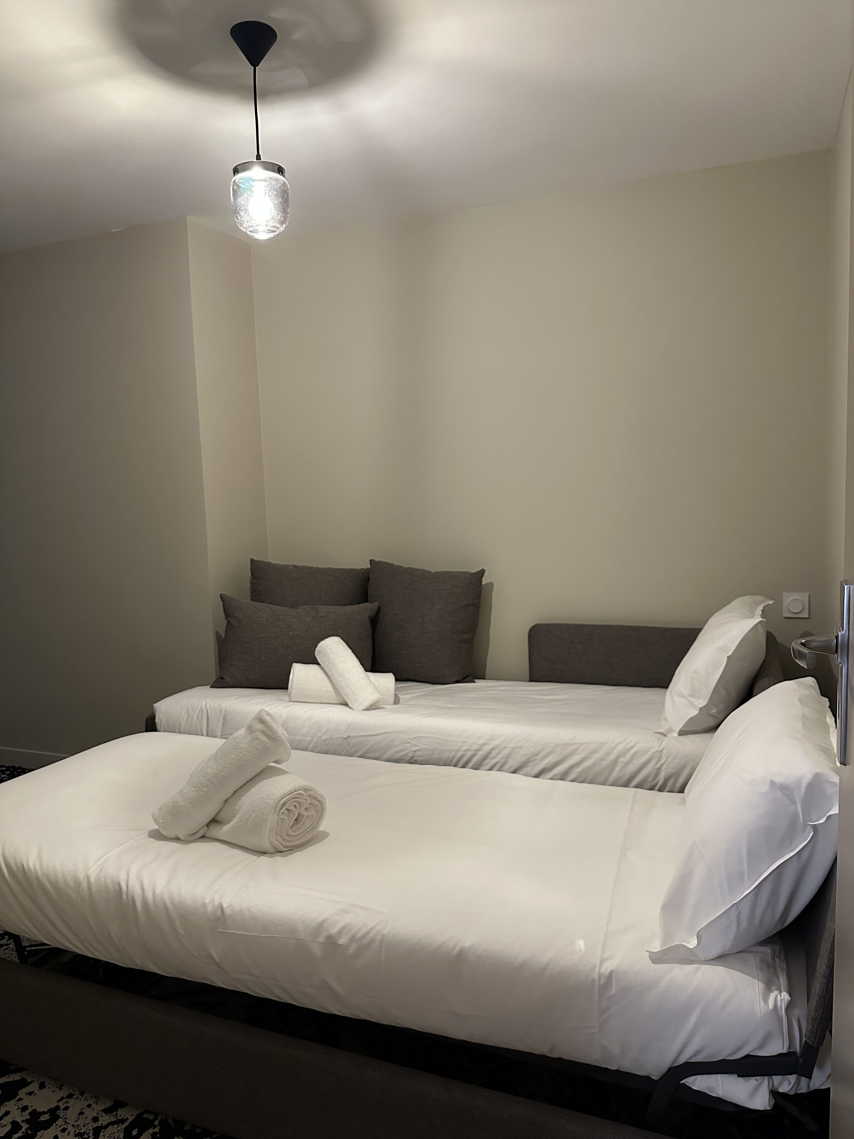 A hotel room with two beds, a hanging light fixture, and a visible power outlet on the wall.