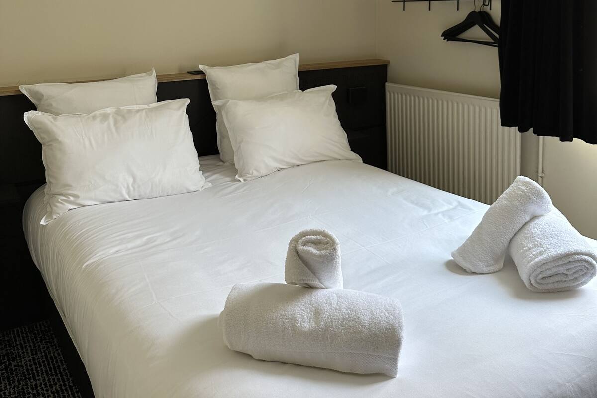 Siisti hotellihuone, jossa on huolitellusti pedattu sänky, valkoiset vuoteeksi, musta pääty, patteri, musta verho ja seinälle kiinnitetty hylly, jonka päällä on maljakko ja henkari.