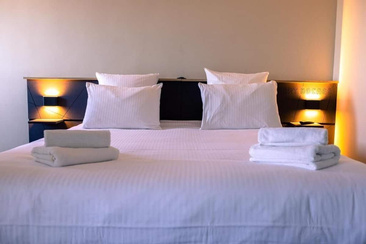 Ein ordentlich bezogenes Bett mit weißen Leinen und Kissen, zwei Nachttischlampen und ein gefaltetes Handtuch auf dem Bett.