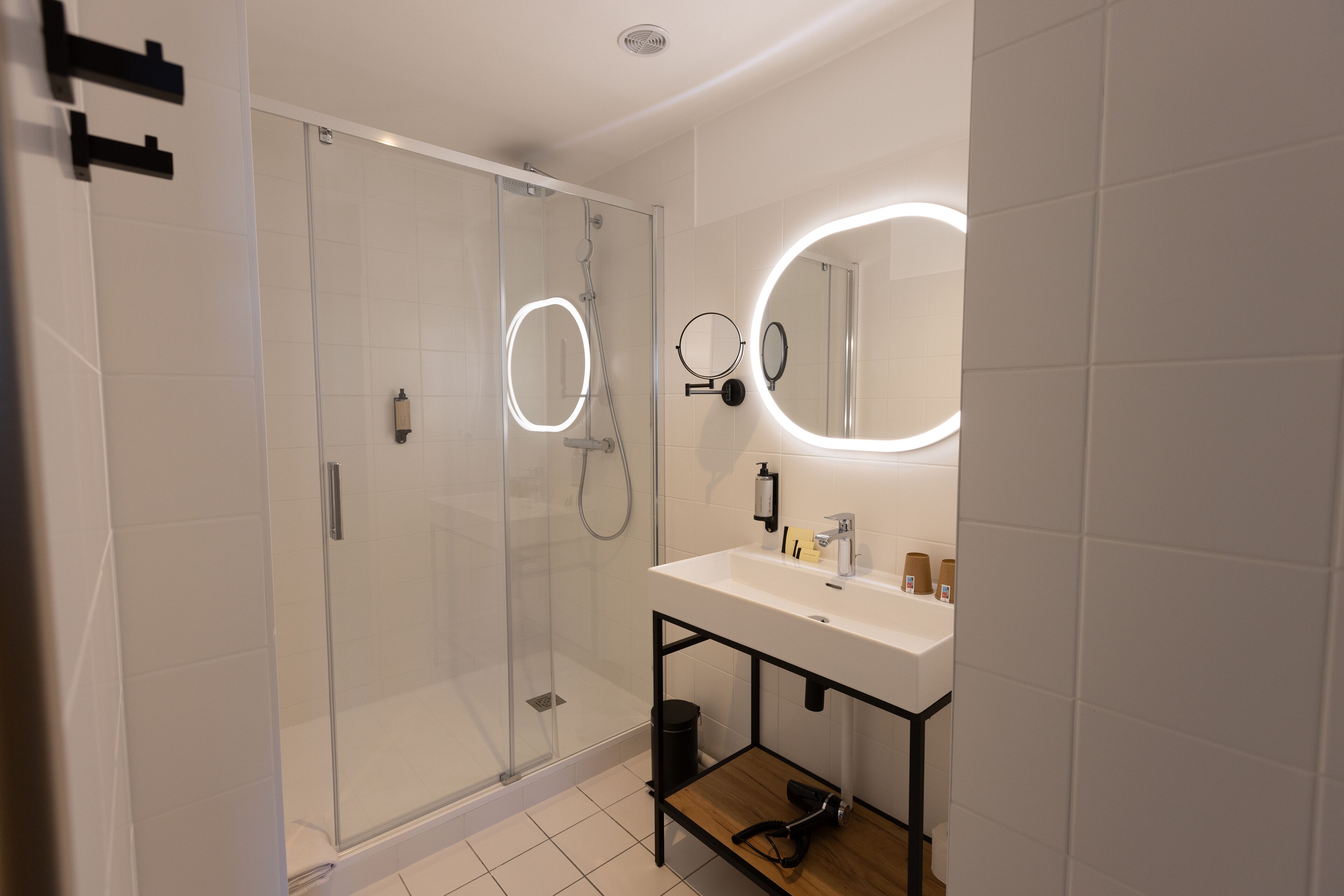 A modern bathroom with a glass-enclosed shower, a white sink with a black frame, and two round mirrors with illuminated edges.